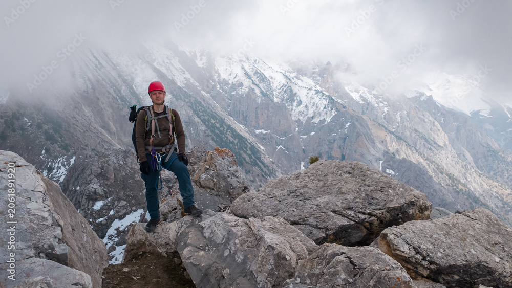 Fototapeta premium A person stands confidently on large rocks in a mountainous area. The sky is cloudy, and snow can be seen on some peaks. This scene shows an outdoor adventure and exploration.