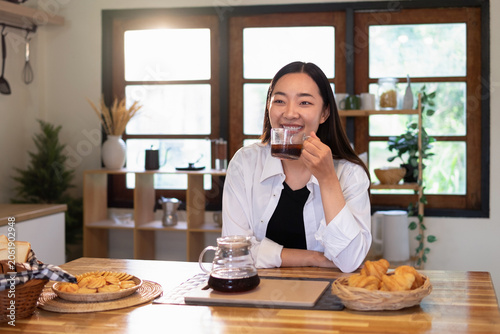 A happy young Asian woman drinks morning coffee at a cozy kitchen table with pastries.