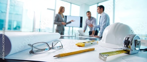 Architectural blueprints with glasses and yellow level on desk against blurred professional team.