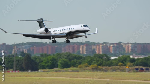 White and black large cabin business jet performs landing at a busy airport with urban cityscape and airport hangar buildings in background, daytime.