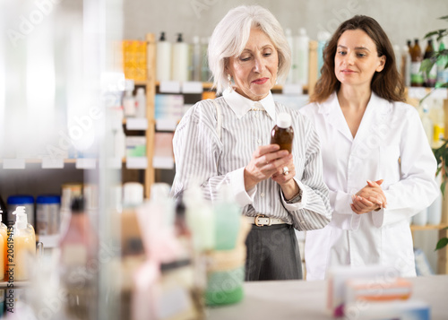 Mature woman choose expectorant syrup, buys product in drug store. Checking for absence of GMOs, age restrictions and recommendations for use. Woman pharmacist offers product, promotes product of
