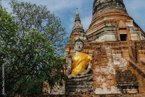 Photography A serene Buddha statue draped in golden cloth sits amidst the ancient ruins of Wat Yai Chai Mongkhon in Ayutthaya, Thailand, a testament to historical Buddhist architecture