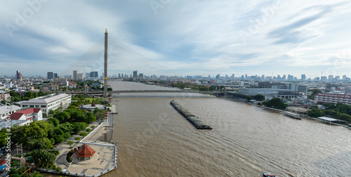 Принти на полотні A barge travels down the Chao Phraya River in Bangkok, Thailand, under the Rama VIII Bridge, showcasing the city's bustling waterway and skyline