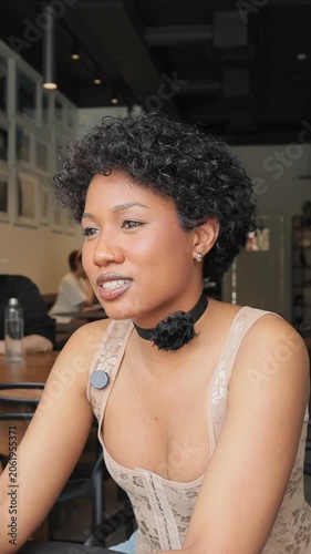 African American woman with curly hair records video blog, black choker sits at table holding white mug. Warm smile, eye contact. Casual cafe setting. Friendly, relaxed, lifestyle portrait.