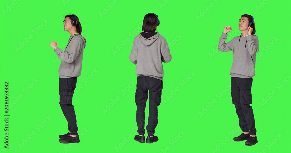 Obraz premium Joyful man does groovy dance moves in studio, feeling funky while he is listening to cool music, with Asian male model smiling and enjoying songs on wireless headset, dancing on green screen.