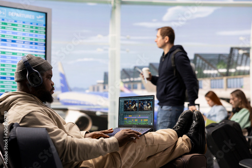 Canvas-taulu Black videographer reviewing film montage and editing in post production, seated in airport lounge before boarding, International travel with nomadic lifestyle, work anywhere concept