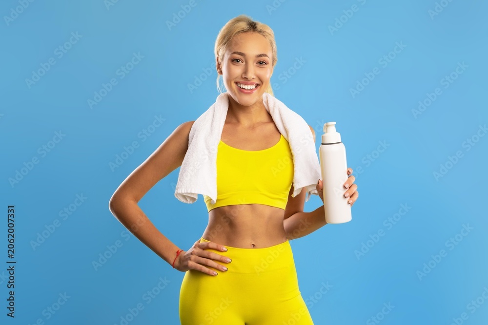 Naklejka premium Smiling Young Woman With Towel Around Neck Holding Water Bottle After Exercising, Standing In Gym Environment, Displaying Happiness And Enthusiasm While Focusing On Fitness Journey.