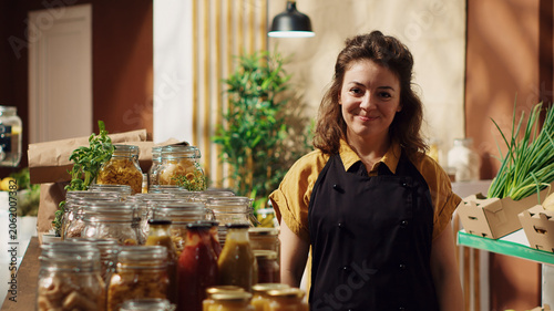 Portrait of smiling zero waste supermarket owner using nonpolluting glass containers for its bulk items to lower climate impact, joyful trader in local grocery shop arranging merchandise.