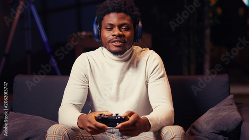 Cuadro en lienzo African American man having online gaming session with friends, talking with them through headphones microphone, gamer playing videogames with teammates on console using gamepad controller