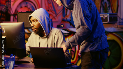 Foto Hacking group new member receiving guidance from knowledgeable rogue programmer acting as his mentor in ghetto abandoned hideaway, master hacker teaches script kiddie how to deploy viruses