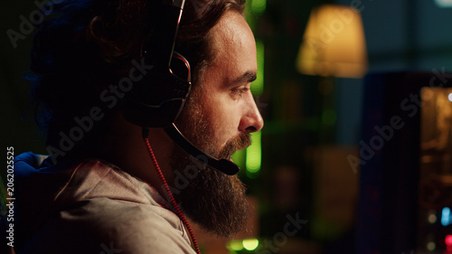 Obraz na płótnie Close-up shot of man relaxing by playing entertaining video game on PC at computer desk late at night, gamer wearing headphones, shooting enemies in online multiplayer first person shooter