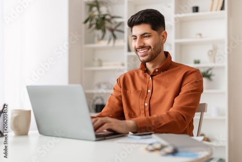 Fotografía Smiling Young Caucasian Man Sitting At Desk Using Laptop In Modern Home Office