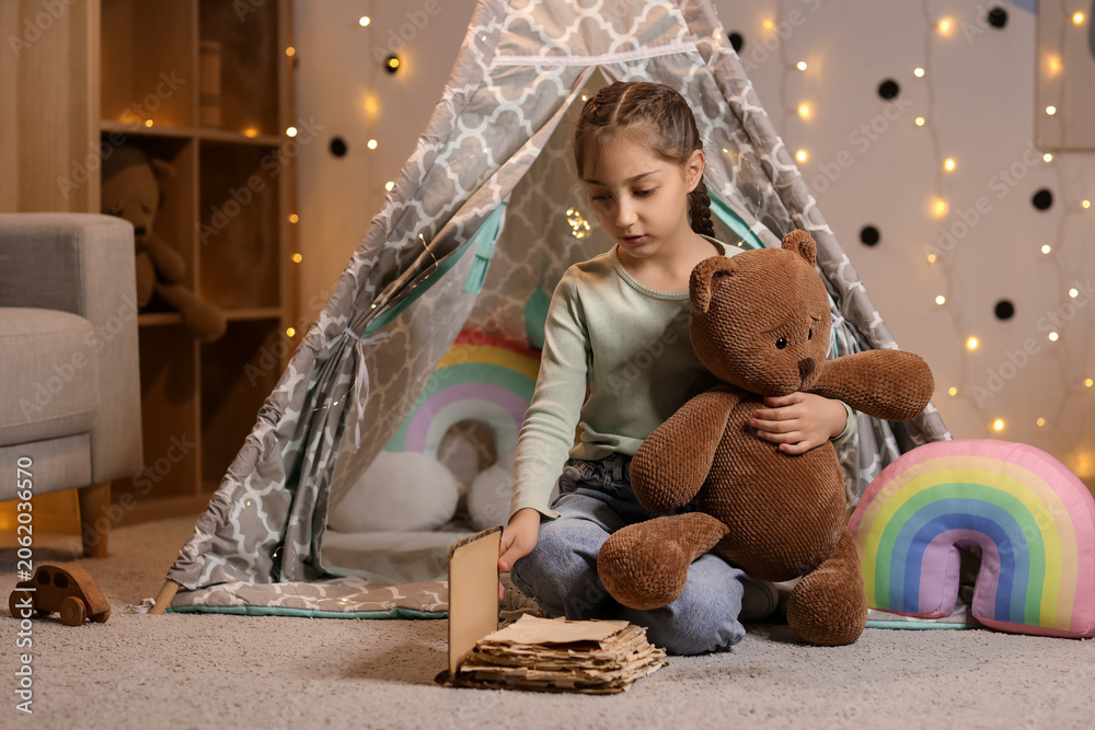 Naklejka premium Cute girl with toy bear reading adventure book at home in evening