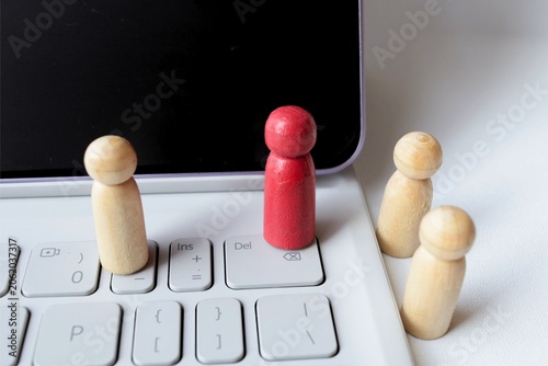 ​A red wooden peg doll standing on a laptop keyboard with natural figures in the background, concept of remote work, digital leadership, and online recruitment.