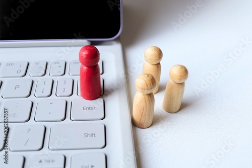 ​A red wooden peg doll standing on a laptop keyboard with natural figures in the background, concept of remote work, digital leadership, and online recruitment.