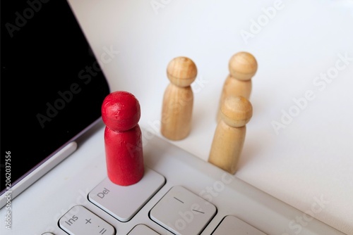 ​A red wooden peg doll standing on a laptop keyboard with natural figures in the background, concept of remote work, digital leadership, and online recruitment.