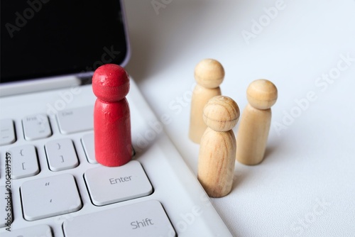 ​A red wooden peg doll standing on a laptop keyboard with natural figures in the background, concept of remote work, digital leadership, and online recruitment.