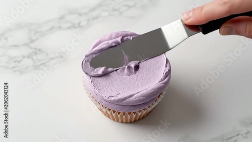 Close-Up Of An Offset Spatula Spreading Pastel Lavender Frosting On A Delicious Cupcake Against A Marble Background