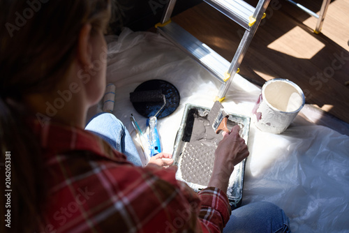 Person holds paintbrush over tray with paint, roller, bucket and ladder nearby on protective sheet.
