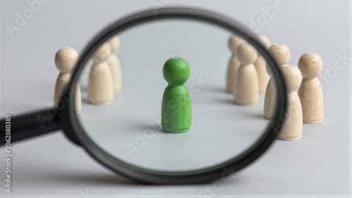 Magnifying glass highlights one green wooden figure among many beige peers.
