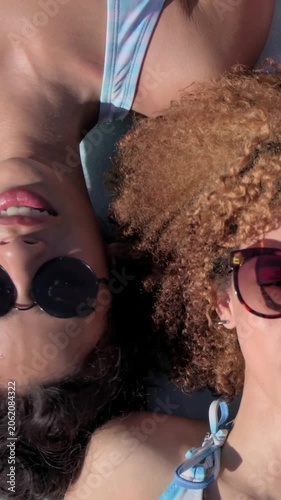 Vertical video: Lying women in bikinis, heads close on dark pad, sunlight causing shades shifting