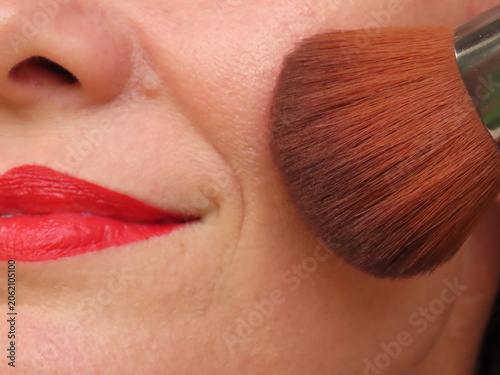 Woman with red lipstick applying makeup with a brush. Woman applying blush using a brush. Woman in her forties doing her beauty routine. Beauty routine concept.