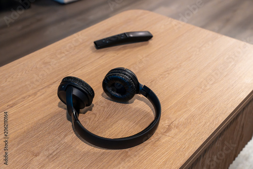 Horizontal photo of black over-ear headphones resting on a light wooden table next to a black TV remote in a minimalist setting