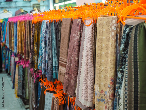 Canvastavla Rows of vibrant, colorful hijabs hang on racks, showcasing batik patterns and a variety of fabrics in the bustling market, inviting a sense of discovery and fashion