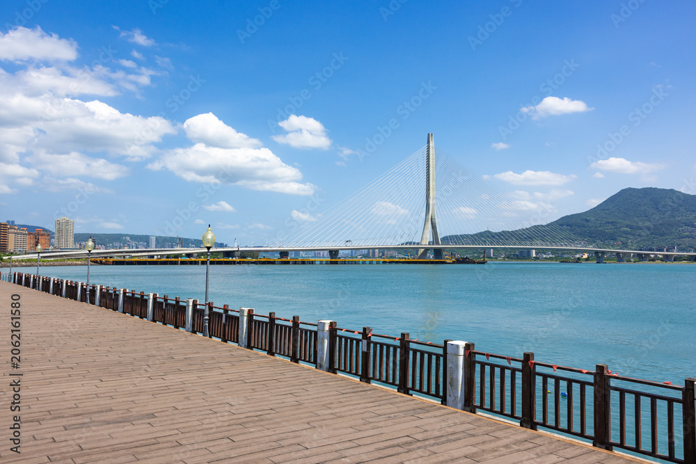 Fototapeta premium Wooden seaside walkway overlooking modern Danjiang Bridge in Tamsui Taiwan