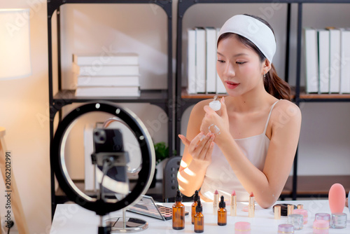 Young asian woman presenting and review cosmetic product with on social media, beauty influencer reviewing skincare cream product on hand with phone, live streaming or broadcast, content creator.