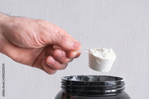 Male hand holds measuring spoon with brown protein on gray background side view.