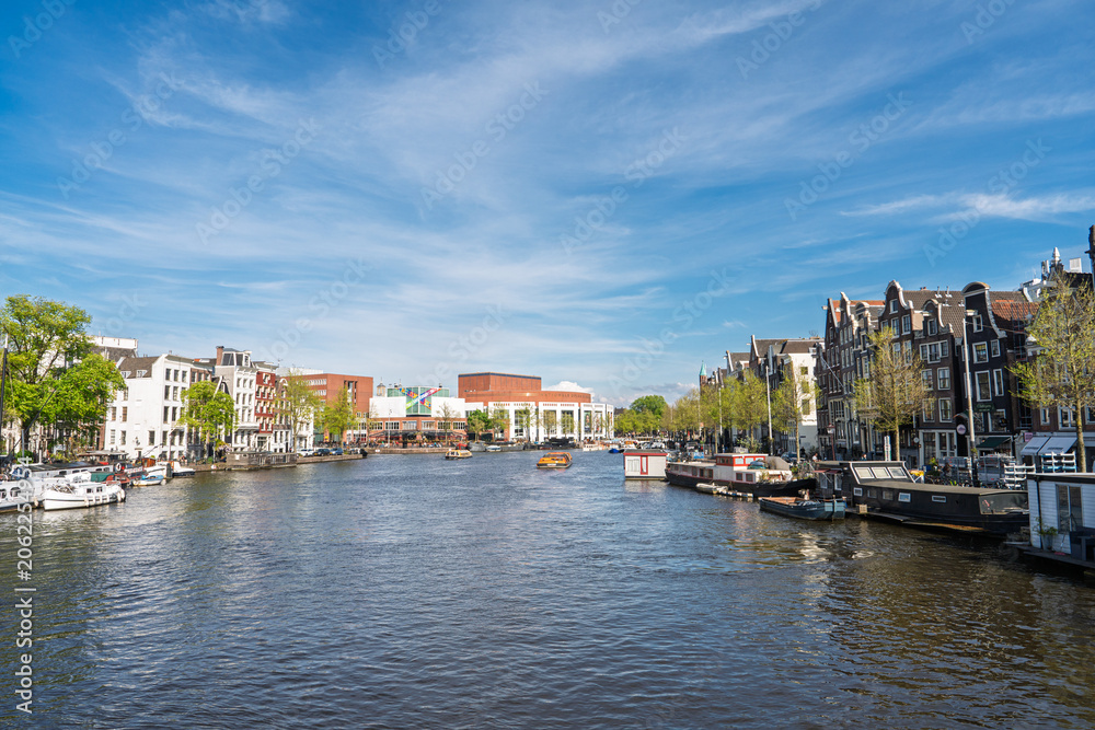 Fototapeta premium A scenic view of the Amstel River featuring historic canal houses and houseboats in Amsterdam, Netherlands. The bright sunny day highlights the classic Dutch urban architecture.