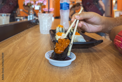 People using chopsticks to pick up sushi roll from ceramic plate with dipping sauce cup inside Japanese restaurant. Woman Customer holding sushi piece with chopsticks over soy sauce cup Japanese Food.