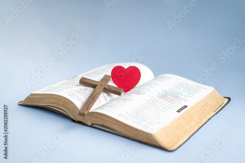 Crucifx cross and red paper heart on top of the Holy Bible. Isolated on light blue background.