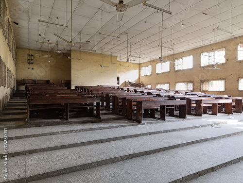 Empty university lecture hall with tiered seating and wooden desks, interior of an aging academic classroom illuminated by natural daylight through barred windows, educational infrastructure and learn