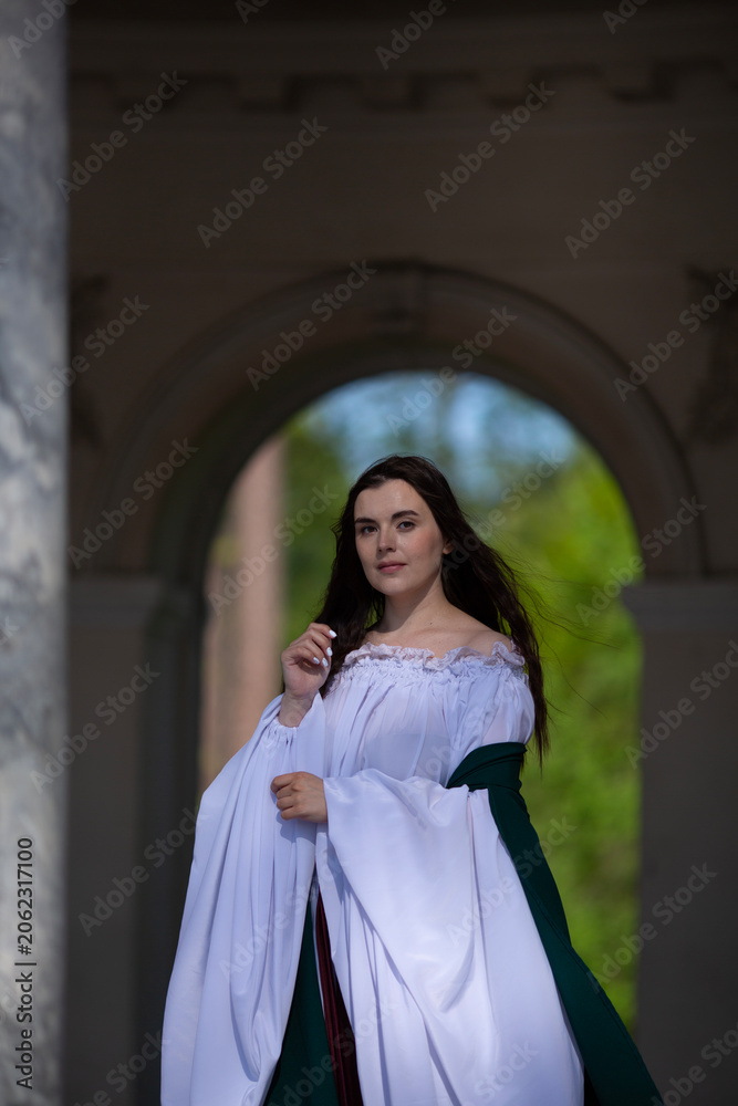 Naklejka premium Vertical photo. Young adult woman with bare shoulder, green white medieval dress stands near arch, marble column of palace, looks at camera. Concept of diverse beauty, Middle Ages, pray, magic, faith