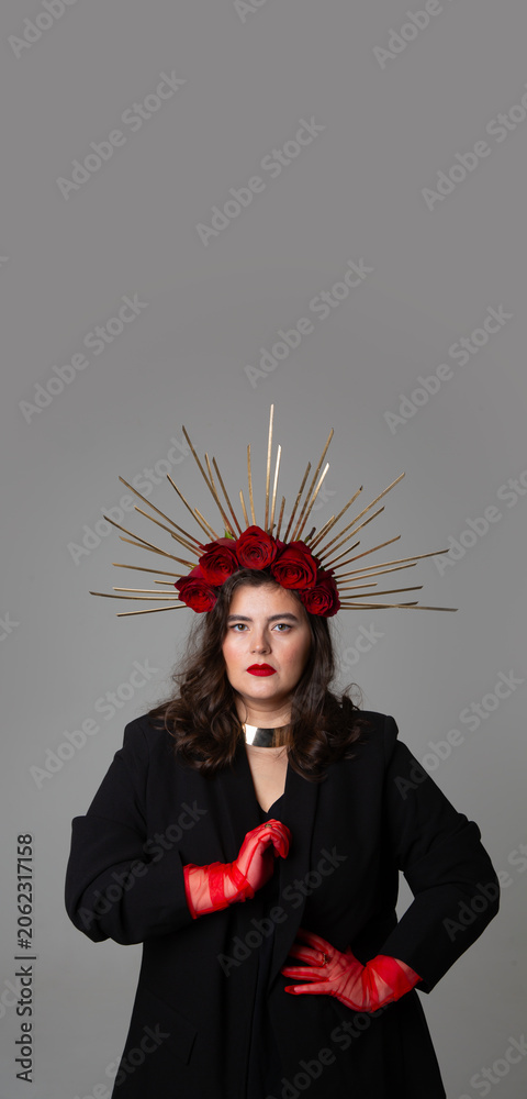 Naklejka premium Vertical photo banner. Young adult white woman with DIY golden halo crown, rose on head, red lips, tulle gloves, black jacket looks at camera on gray. Diverse beauty, pray, faith, fantasy. Copy space