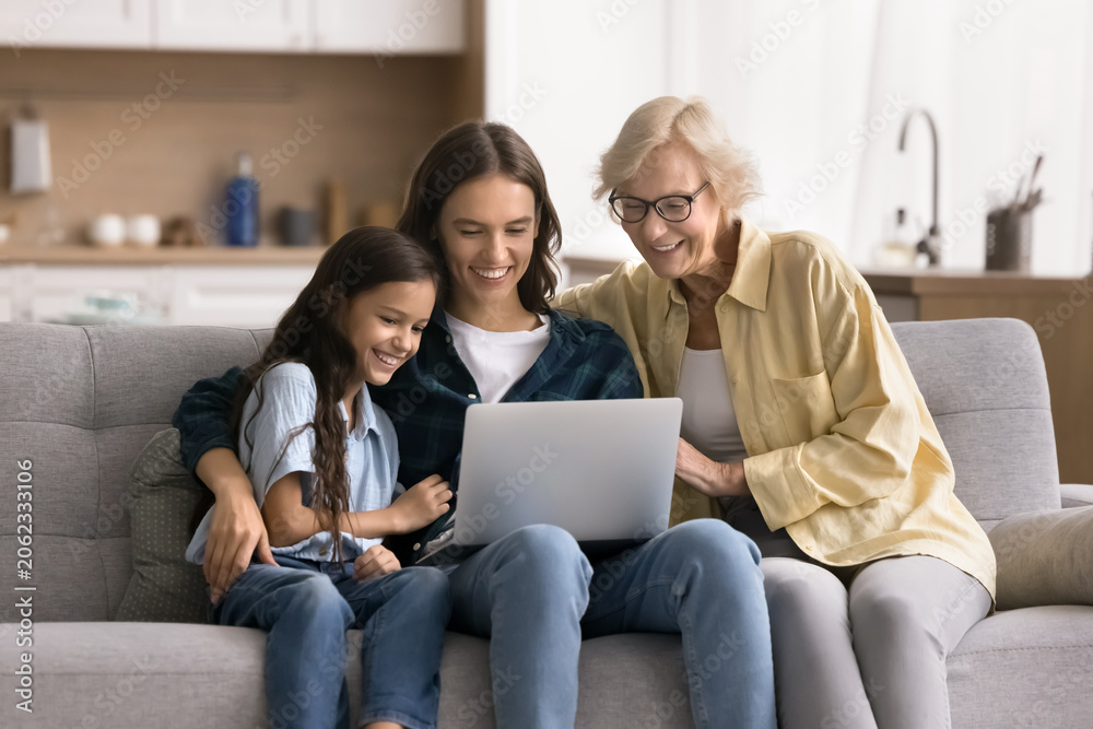 © fizkes - Little girl spend weekend with mom and older granny watching movie using on-line digital streaming subscription services, buying goods and e-services via marketplace website. Tech, fun, family leisure