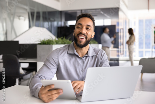 Tableau sur toile Happy businessman sitting at desk with tablet and open laptop, reviewing information and results on screen, laughing, enjoying good news, successful project completion