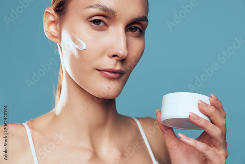 Foto Woman applying white face cream, holding a small jar, close up on blue backgroun