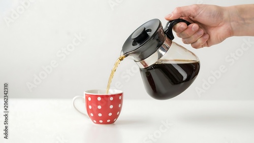 A person pours coffee from a glass pot into a red polka-dot cup on a table indoors.