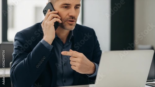 Man on Phone and Laptop A focused businessman in a suit speaks on his mobile phone while gesturing towards an open laptop engaged in a serious discussion