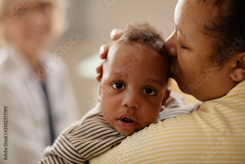 Portrait of Black mother holding infant during pediatrician appointment, comforting child while doctor waiting in background. Ideal for pediatric care, parenting support, child health marketing