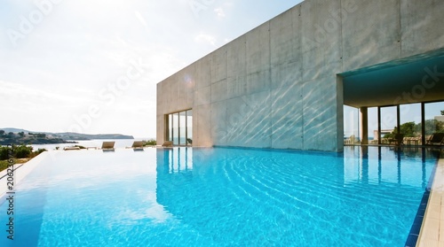 Infinity pool at modern luxury villa overlooking mediterranean sea. Minimalist concrete architecture with water reflection. Summer vacation destination for peaceful travel and relaxation.