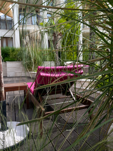 Vertical photo of a luxury wooden chaise lounge with a vibrant pink cushion on a sun deck framed by long green ornamental grass blades with a resort hotel building in the background