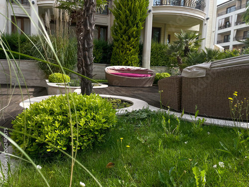 Horizontal photo of a luxury resort courtyard garden featuring lush green lawns, trimmed bushes, a palm tree trunk, and outdoor wicker patio furniture on a wooden deck by a hotel building
