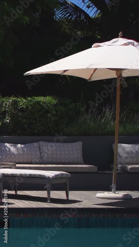 Vertical video: Starting camera panning, beige umbrella shifting right over pool with bird perching