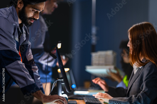 Tableau sur toile South Asian male engineer focuses on a monitor while a female professional and a woman in a yellow safety vest confer in the background