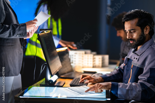 Focused South Asian engineer types at a desktop workstation while a female colleague holds a report and a safety vest professional stands in the background.