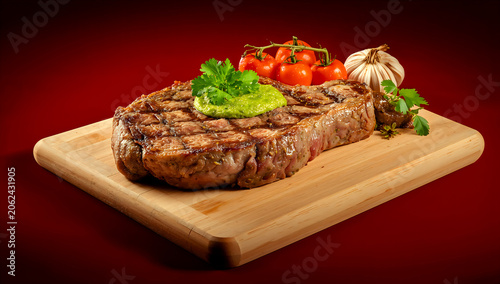 Juicy Ribeye Steak with Chimichurri, Cherry Tomatoes and Roasted Garlic on Red Background BBQ Braai Scene
